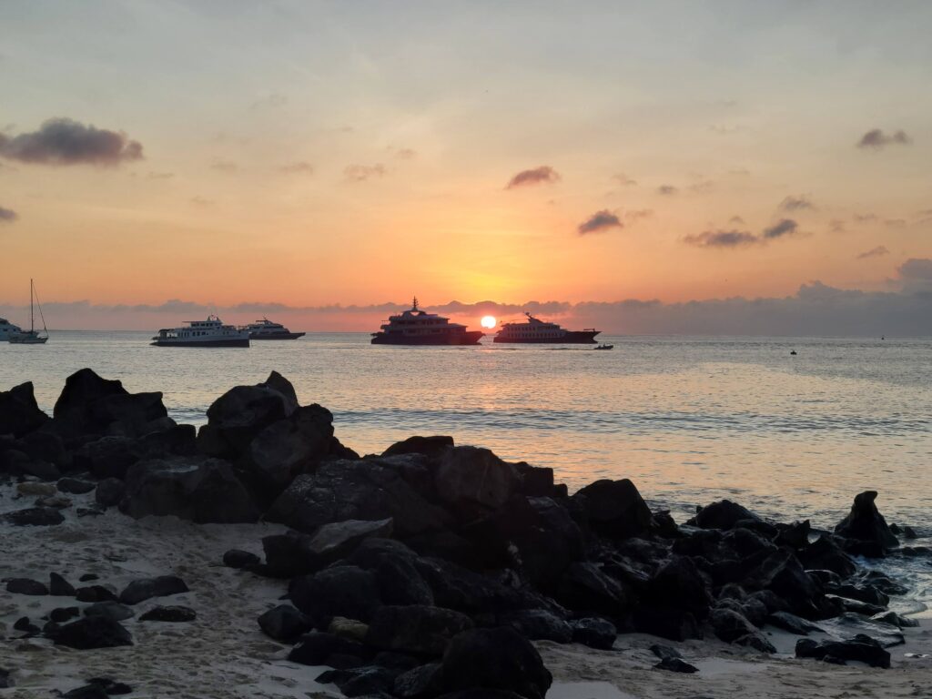 ocean sunset viewed from sandy beach with lava rock and galapagos cruise boats