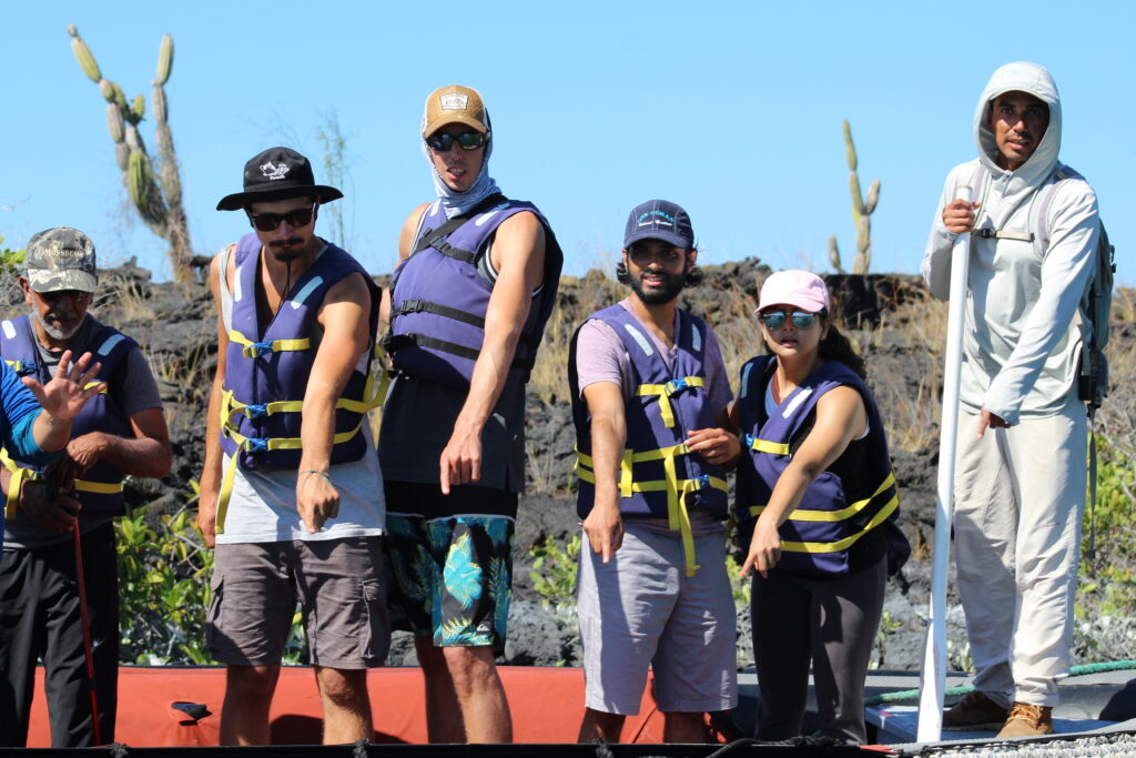 tourists in life jackets with a Galapagos national park guide stand on a dinghy pointing at the water